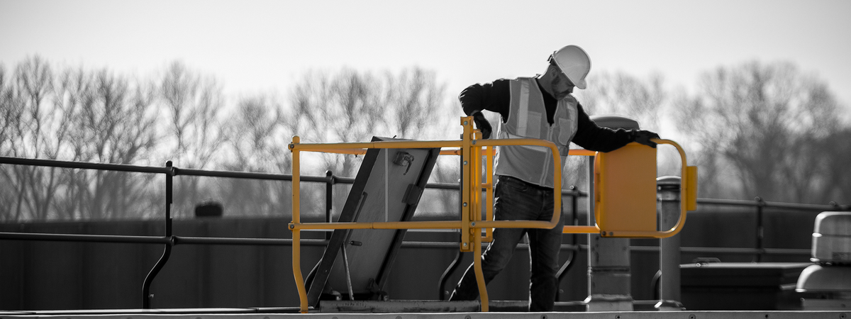 Worker using a roof hatch guardrail gate, with the yellow guardrail highlighted against a black-and-white rooftop scene