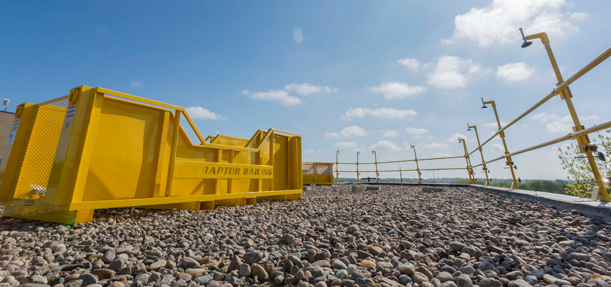 Yellow Raptor Rail guardrail crate and railing system installed along a gravel commercial rooftop under a blue sky